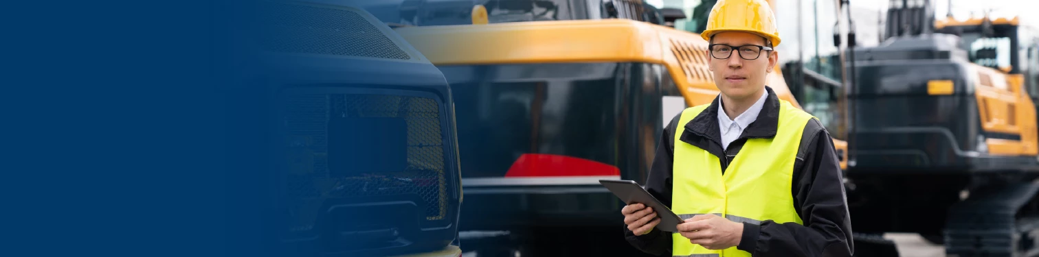 Man in a suit and hardhat holding a clipboard in front of construction equipment.