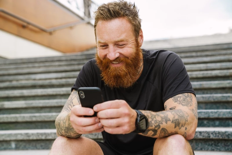 Close up image of a man smiling while on his phone and sitting on a park bench