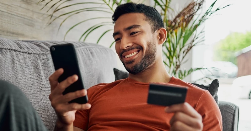 Image of man sitting on his couch with his phone in one hand and his card in another
