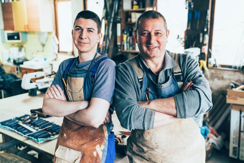 A business owner stands and smiles next to his younger employee and considers his business banking and succession options