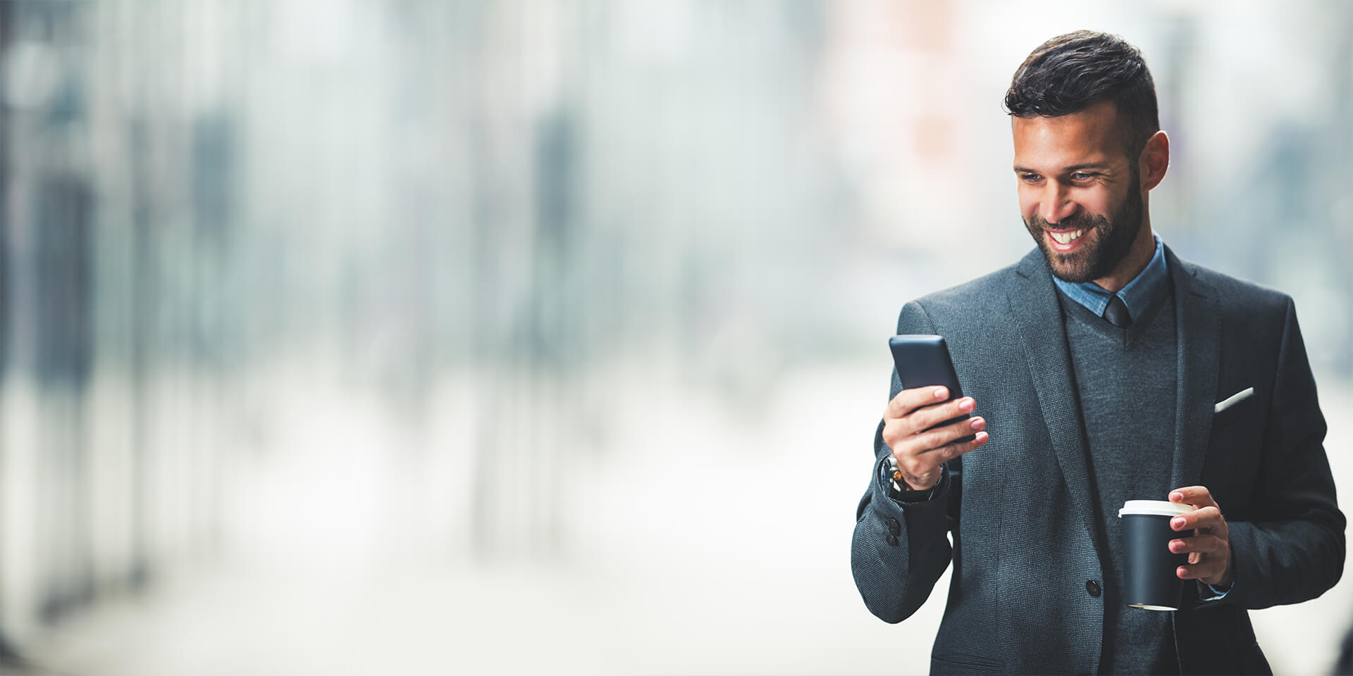 A businessman is holding a cup of coffee and confidently looking at his business checking account on his mobile device
